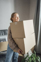 Smiling young woman carrying cardboard boxes in new apartment - VPIF05213