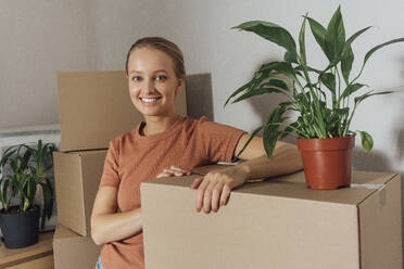 Lächelnde Frau mit Topfpflanze, die sich zu Hause auf Kartons stützt - VPIF05230