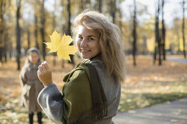 Lächelnde Frau hält Herbstblatt in öffentlichem Park - LLUF00327
