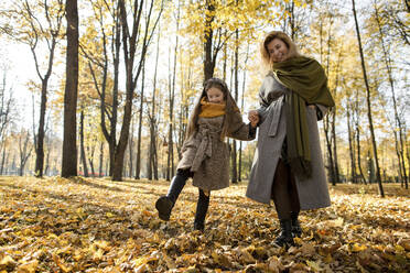 Smiling woman holding hand of daughter kicking autumn leaves in park - LLUF00334