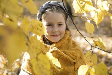 Lächelndes Mädchen mit Wollschal schaut durch einen Zweig in einem herbstlichen Park - LLUF00336