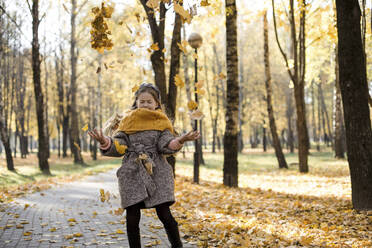 Carefree girl throwing autumn leaves on walkway in park - LLUF00337