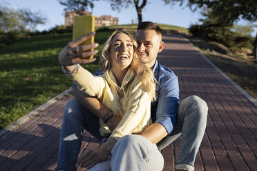 Smiling girlfriend taking selfie with boyfriend through mobile phone at sunset - JCCMF04504