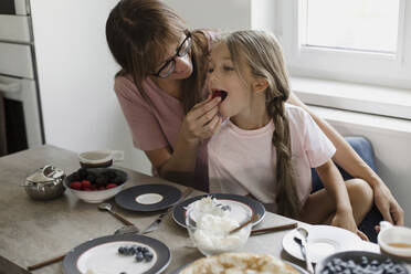 Mother feeding berry to daughter at table - LLUF00352