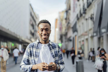 Smiling man wearing plaid shirt holding smart phone in city - PGF00918