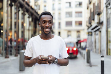 Smiling man holding smart phone at city street - PGF00926