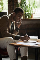 Lächelnder junger Mann, der in einem Café mit seinem Smartphone telefoniert und in ein Notizbuch schreibt - DSHF00074