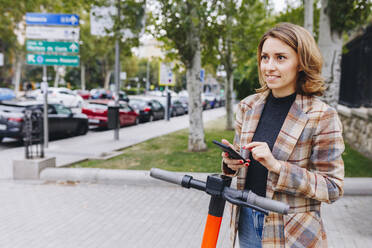 Smiling freelancer with mobile phone and electric push scooter on footpath - MRRF01681