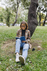 Smiling freelancer reading book in front of tree in park - MRRF01689