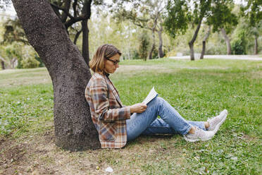 Businesswoman leaning on tree trunk and reading book in park - MRRF01690