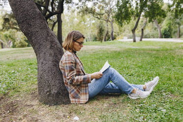 Geschäftsfrau lehnt an einem Baumstamm und liest ein Buch im Park - MRRF01690