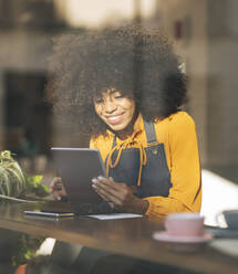 Geschäftsfrau mit Schürze benutzt Tablet-PC am Fenster eines Cafés - JCCMF04530
