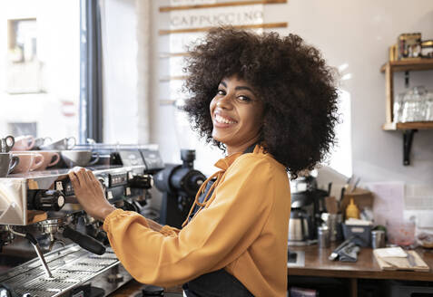 Lächelnde Kellnerin mit Afrofrisur an der Kaffeemaschine stehend - JCCMF04542