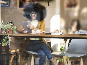 Lächelnde Frau mit Afrofrisur, die einen Tablet-PC am Fenster eines Cafés benutzt - JCCMF04564