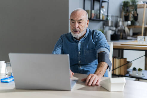 Älterer Mann mit Blutdruckmessgerät, der zu Hause einen Arzt am Laptop konsultiert - GIOF14121