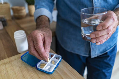 Mann hält ein Glas Wasser und nimmt Medikamente aus einer Pillenschachtel zu Hause - GIOF14147