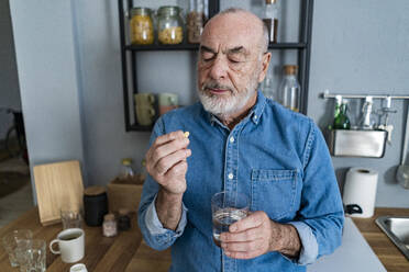 Älterer Mann hält ein Glas Wasser und betrachtet ein Medikament in der Küche - GIOF14148