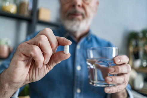 Senior man holding glass of water and pill at home - GIOF14152