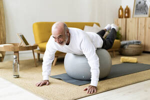 Active senior man exercising on fitness ball at home - GIOF14175