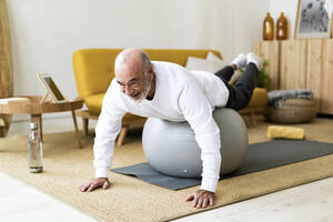 Aktiver älterer Mann, der zu Hause auf einem Fitnessball trainiert - GIOF14175