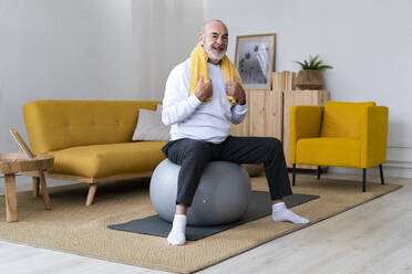 Älterer Mann mit Serviette um den Hals sitzt zu Hause auf einem Fitnessball - GIOF14178