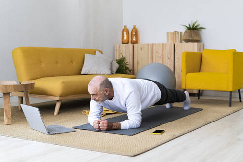 Älterer Mann mit Laptop übt die Plank-Pose auf einer Übungsmatte - GIOF14190