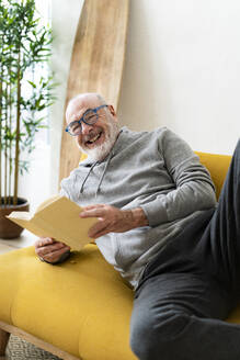 Lächelnder Mann mit Buch entspannt auf Sofa im Wohnzimmer - GIOF14198