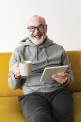Älterer Mann lächelt, während er mit Kaffeetasse und Tablet-PC zu Hause sitzt - GIOF14204