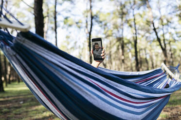 Woman's hand holding smart phone on video call relaxing in hammock at park - JCCMF04574