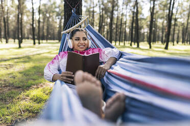 Lächelnde Frau mit Buch in Hängematte liegend und Musik hörend im Park - JCCMF04577