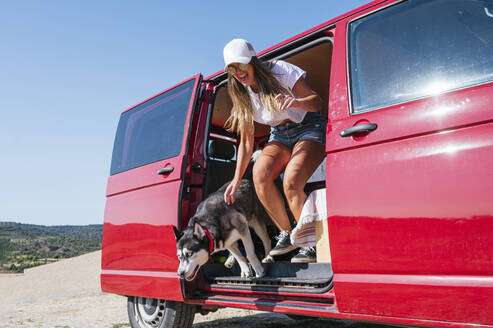 Fröhliche Frau springt mit Hund aus rotem Lieferwagen an sonnigem Tag - JCMF02243