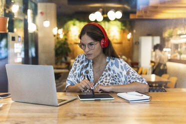 Businesswoman wearing headphones using digital tablet in coffee shop - PNAF02554