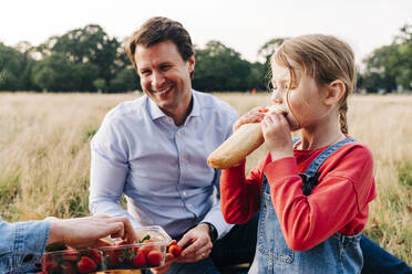 Girl eating loaf of bread at picnic with family in park - ASGF01675