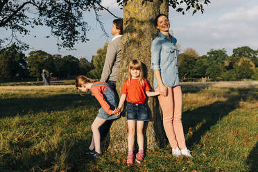 Familie hält sich an den Händen und steht um einen Baum im Park - ASGF01761