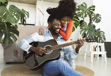 Young woman embracing boyfriend playing guitar in living room - JCCMF04591