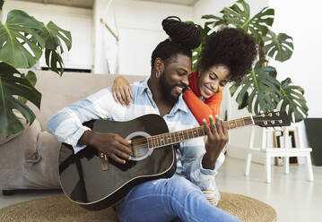 Curly haired woman embracing boyfriend playing guitar at home - JCCMF04592