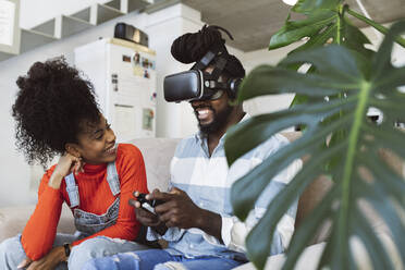 Smiling woman looking at boyfriend with virtual reality simulator playing game in living room - JCCMF04595