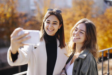 Teenage girl taking selfie with friend through smart phone in autumn park - DAWF02294