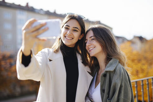 Smiling teenage girl wearing coat taking selfie with friend at park - DAWF02295