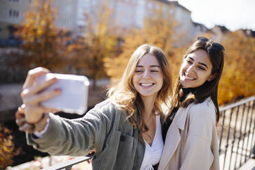 Glückliches Teenager-Mädchen nimmt Selfie durch Smartphone mit Freund am Geländer - DAWF02296