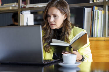 Businesswoman with diary using laptop in coffee shop - IFRF01230