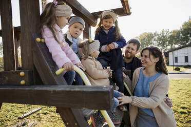 Happy parents looking at children sitting on outdoor play equipment - LLUF00368