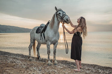 Frau, die auf einem weißen Pferd am Ufer entlang reitet - CAVF95289