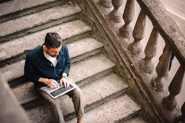 Man using laptop computer sitting on steps in city - CAVF95292