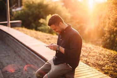Glücklicher Mann mit Smartphone auf einer Bank sitzend bei Sonnenuntergang - CAVF95294