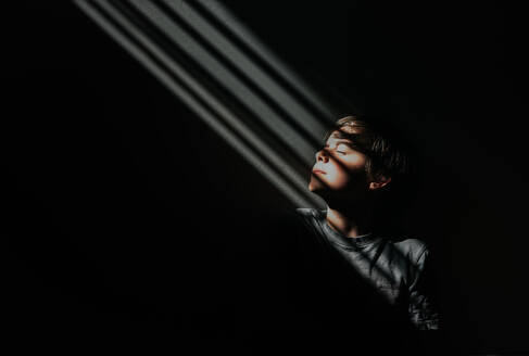 Young boy sitting in a patch of patterned light in a dark room. - CAVF95298