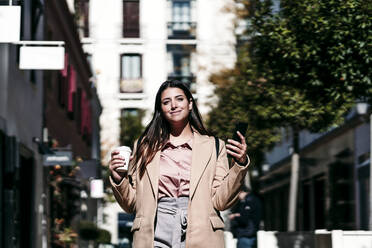 Lächelnde Geschäftsfrau mit Einwegbecher und Mobiltelefon auf einer Straße in der Stadt - EBBF04813