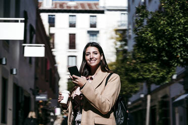 Smiling businesswoman holding smart phone in city - EBBF04816