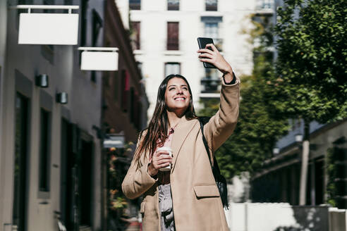 Lächelnde Geschäftsfrau, die ein Selfie mit ihrem Smartphone an einem sonnigen Tag macht - EBBF04817