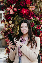 Junge Geschäftsfrau mit Handy in der Hand vor Blumen - EBBF04822